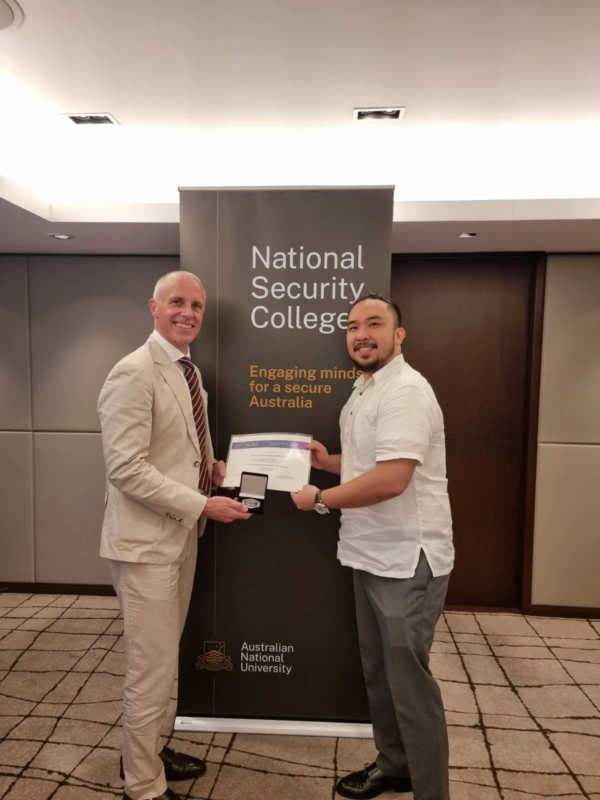 Rainier Milanes standing with another adult beside an Australian National University National Security College banner, holding a certificate.