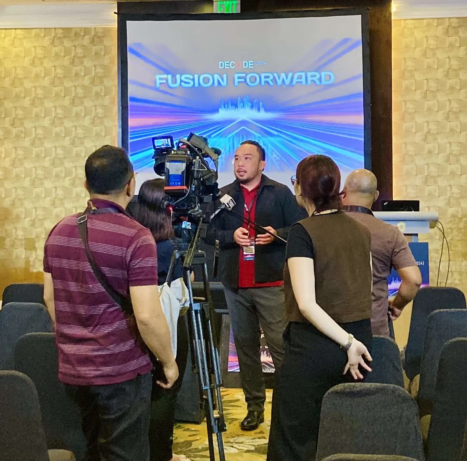 Rainier Milanes speaking during a media interview in front of a Decode 2024 event screen.